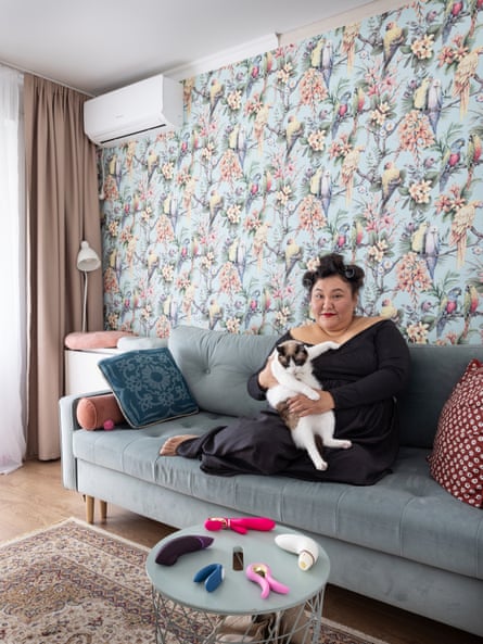 Portrait of Gulim sitting with her legs underneath her on a couch while cuddling a tabby cat. On a small table in front of her are her sex toys