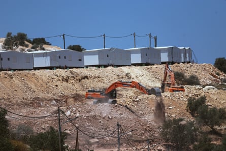 Israeli bulldozers prepare land on a hillside. Temporary cabin structures are lined up above the ridge.
