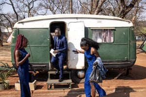 A man wearing a dark blue three-piece suit and holding a white document steps out of the caravan, flanked by two women in dark blue waiting outside