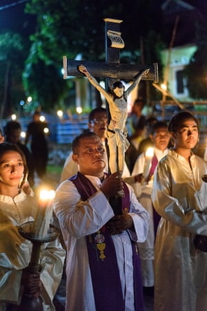 A priest carries a crucifix while about him, members of a choir wait for the procession to continue through the streets.