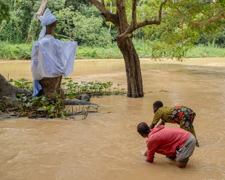 Two African girls up to their knees in a brown river near a large crude wooden sculpture of a figure draped in a white cloth