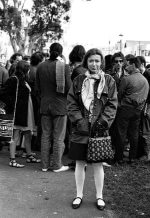 Joan Didion, wearing white tights and Mary Jane flats, stands near a large crowd in San Francisco's Golden Gate park in this black and white image from 1967.