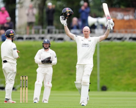 The Lancashire batter Luke Wells raises his bat and takes off his helmet to celebrate scoring a hundred in the 2025 County Championship against Kent at Blackpool