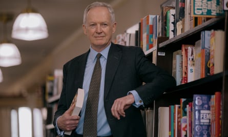 An older white man wearing a suit and tie leans one elbow on a bookshelf, holding a book curled to his chest with the other hand.
