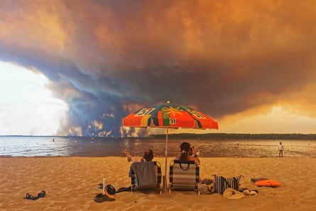 A couple sit on a beach as clouds of smoke rise into the sky across the water