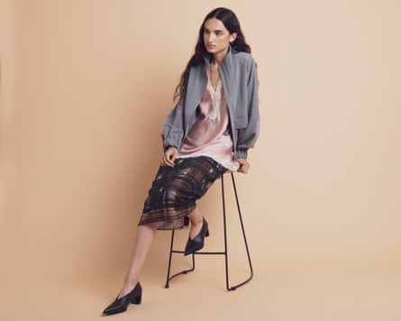 A model sitting ona stool wearing a dark patterned midi skirt, a lacy camisole and a grey jacket