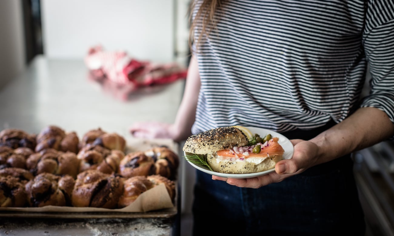 A women holds up a plate on which there is a salmon and cream cheese bagel; behind her is a table strewn with fresh bagels, from Fine Bagels in Berlin.
