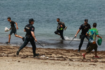Two police officers at shoreline with batons drawn confront a person in green shirt
