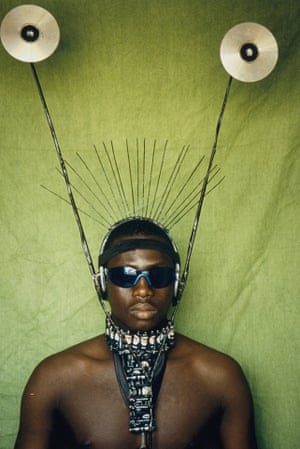 A black model wears a necklace made from batteries and CD antennae. He is bare chested and wearing wrap-around sunglasses.