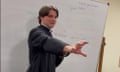 A young white man with shaggy brown hair in a black robe and white clerical collar stands in front of a white board gesturing with one hand.