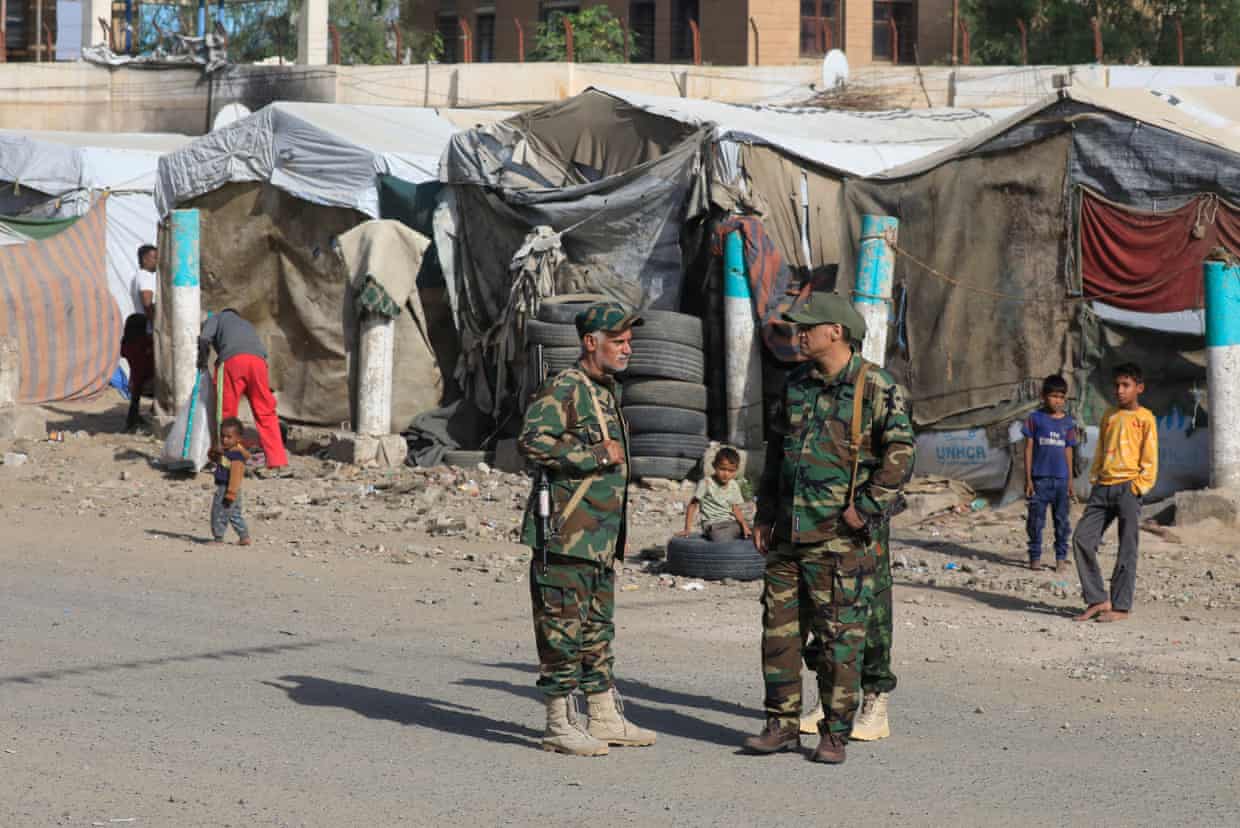 Two men in camouflage, carrying guns, stand in front of a line of tents as children look on.