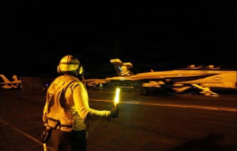 A crew member in a yellow vest and helmet holds a light wand on a carrier deck as a jet takes off at night