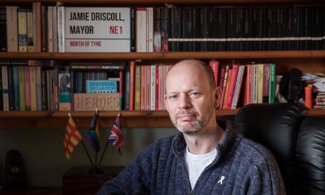 Jamie Driscoll in front of a bookshelf at his home with a sign saying "Jamie Driscoll, mayor"