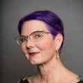 A woman with purple hair and cat’s-eye glasses smiles in a portrait photo.