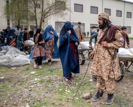 Man in camouflage uniform with gun over shoulder holding long stick next to bags of food, beside column of women in burqas
