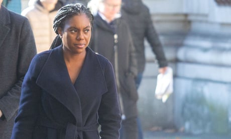 Kemi Badenoch photographed outdoors wearing a dark blue coat with tied belt; she is walking in bright but wintry sunlight past a stone building with blurred people moving behind her