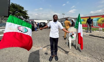 A young man in a white T-shirt and black jeans waves a green, white and red union flag as he stands in front of the airport site. Another flag and several other people, and a pile of white plastic chains, are seen behind him.