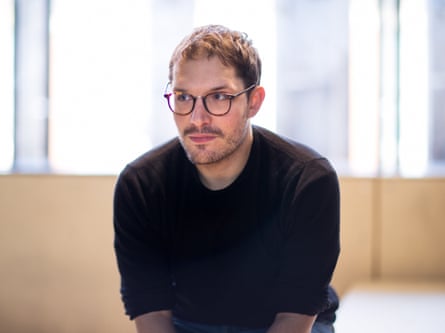 A man in glasses and a dark jumper is shown sitting down.