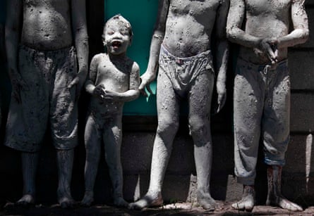 A little boy covered in mud smiles as he stands with three other boys also plastered in grey mud