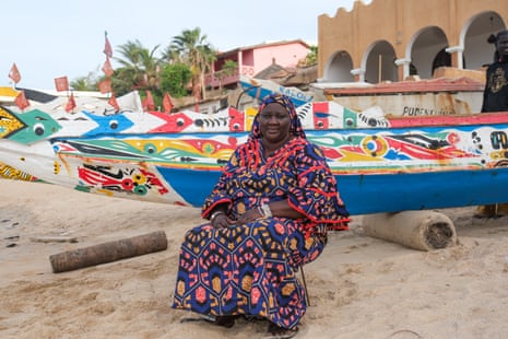 A woman sits on a chair on a beach with colourful boats behind her