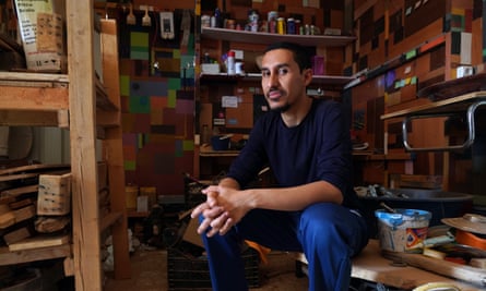 Mohamed Sleiman Labat, a bearded young man, sits smiling reflectively with his hands together among shelves of pots and sticky notes
