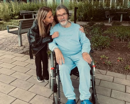 Horace Mason, 74, sits in a wheelchair in a garden. He wears a light blue tracksuit and turquoise Croc-style rubber clogs, and has a grey beard. A woman with long, highlighted blond hair leans over him.