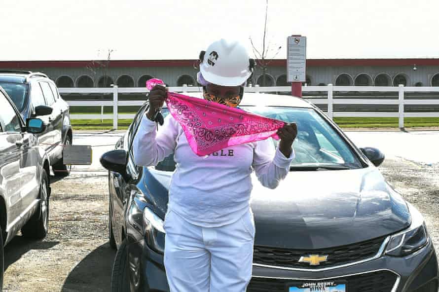 A Black woman wearing a hard hat stands next to a car in a parking lot, adding a second bandana to the one she is already wearing.