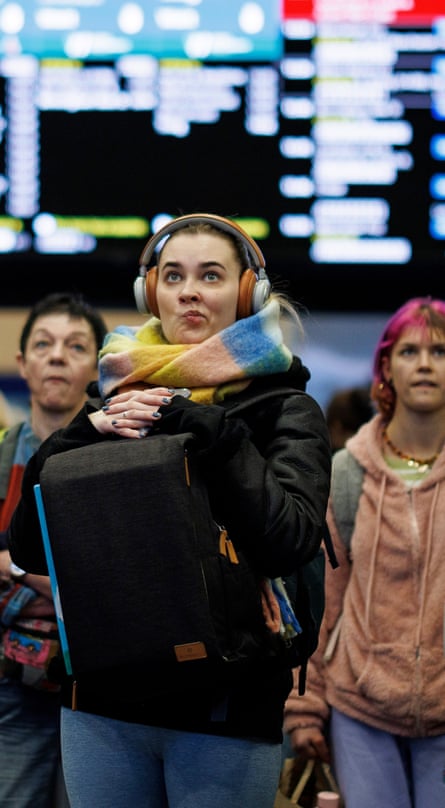 A woman with earphones and a scarf rests her hands on her backpack in front of her, and scrunches her mouth sceptically, with an electronic train timetable behind her