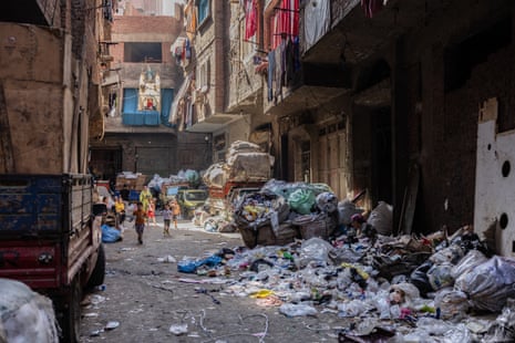Children play on a street lined with piles of rubbish. Sacks of rubbish are also piled up around parked cars