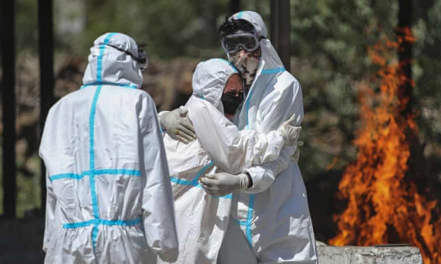 Two people in white hazmat suits hug in front of a funeral pyre, as a third looks on