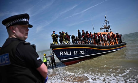A British police officer stands guard on the beach of Dungeness, on the southeast coast of England, on June 15, 2022, as Royal National Lifeboat Institution's (RNLI) members of staff help migrants to disembark from one of their lifeboat after they were picked up at sea while attempting to cross the English Channel.