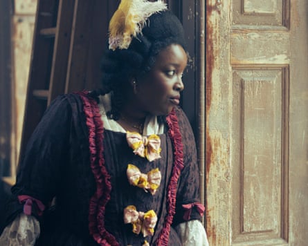 Lolly Adefope as Kitty Higham, in an ornate historical dress and feathered headpiece, gazes through a doorway