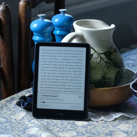 A Kindle e-reader displaying text stands propped against a jug, surrounded by blue pepper mills on a table