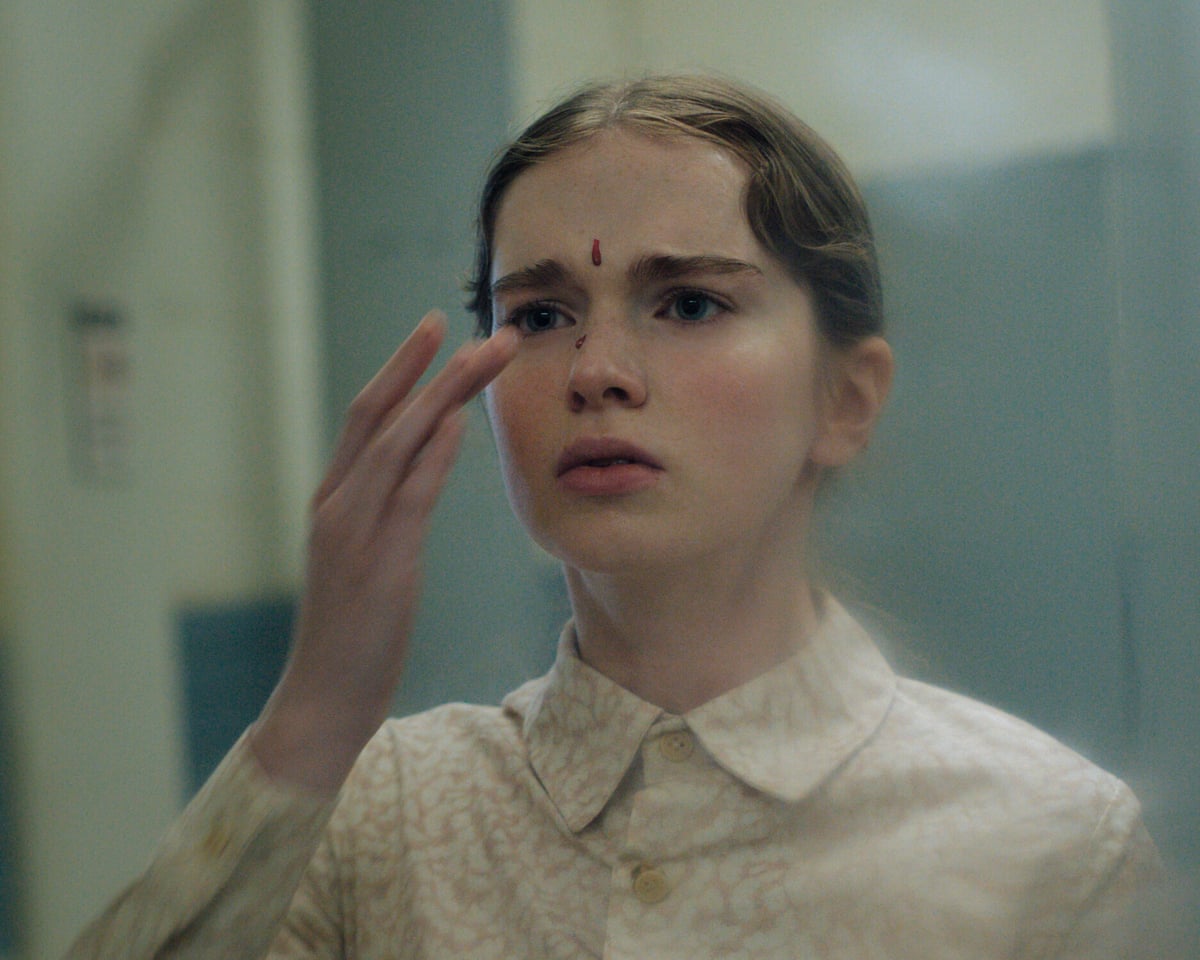 The actress is standing in a corridor, her hand held to her face, which is flecked with blood