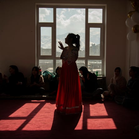 An unveiled woman stands in silhouette against a window; veiled women can be seen sitting around her. The city of Kabul and mountains beyond can be seen through the window