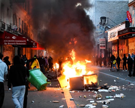 In Paris, a burning bin blazes in a city street as masked figures gather amid smoke and litter.