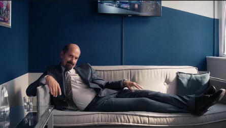 he is reclining on a grey sofa with his feet up. He wears a dark grey suit, white shirt and tie.