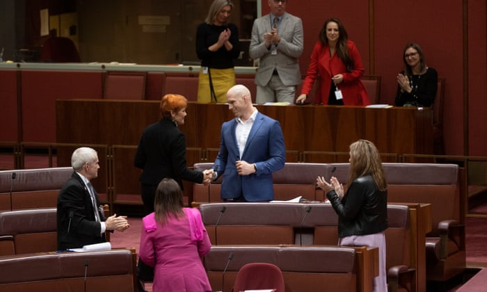 David Pocock shakes hands with Pauline Hanson while standing in his Senate seat. Other senators look towards him and clap