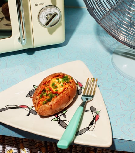 A retro-looking kitchen counter with a microwave, a fan and a triangular, patterned plate with a filled baked potato and a knife on it