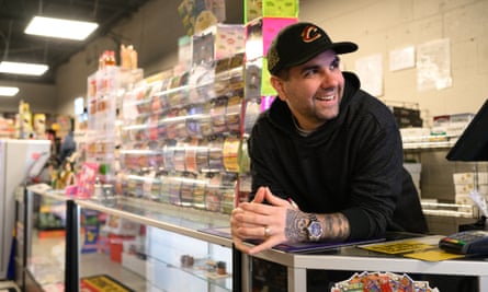 Brian Acierno, with tattoos on his forearm and wearing a baseball cap and hoodie, laughs as he leans on the counter of Cigarettes 4 Less