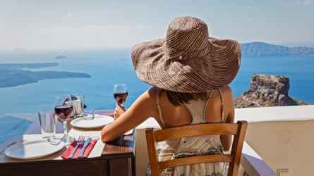 A person in summer dress and large sun hat drinks red wine while looking at the sea from a white terrace