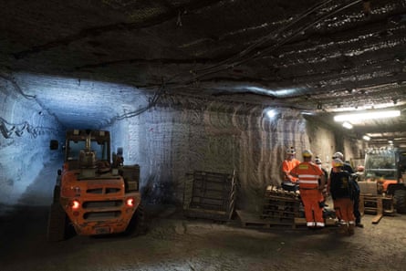 A handful of people wearing orange safety gear and hard hats gather around a stack of pallets at a fork in a tunnel near two forklift trucks.