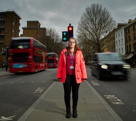 Transport for London’s Katie Willis standing on a paved area in the middle of a London road, traffic lights behind her, two red buses heading away from her one side and a London taxi coming towards her with its lights on