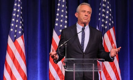 A middle-aged white man in a suit and tie speaks with both palms raised on a stage, standing in front of three American flags on flagpoles.