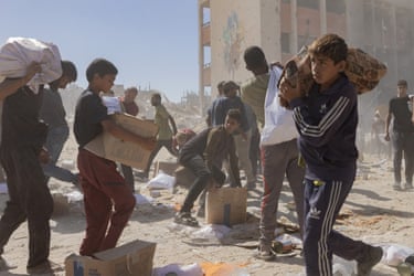 Boys carry boxes and sacks of provisions with the rubble of destroyed buildings in the background