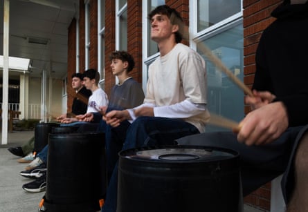 Five young men sit on buckets holding drumsticks in front of a brick building with large windows
