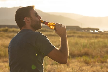 A man drinks beer from a bottle in a large area of grassland with mountains in the distance