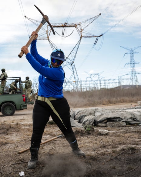 A person in a blue shirt swings a pickaxe at dirt, with armed personnel and a military vehicle behind them