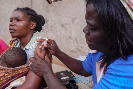 A woman injects another woman, who is holding a baby, in the upper arm