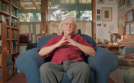 A man with grey hair, a red shirt and grey trousers sitting in a blue armchair with a bookshelf and pictures behind him